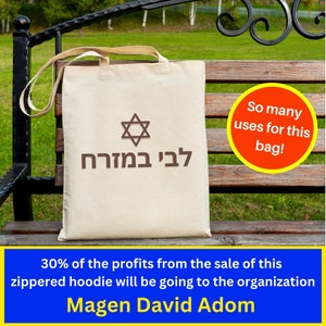 May include: A beige tote bag with brown Hebrew text that reads "L'vi B'mazrach" and a brown Star of David. The bag is sitting on a wooden bench with a green grassy background. A red circle with yellow text reads "So many uses for this bag!"  30% of the profits from the sale of this zippered hoodie will be going to the organization Magen David Adom.