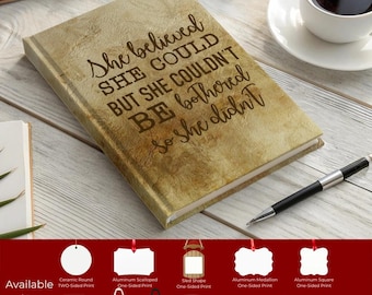 May include: A hardcover journal with a textured, tan cover. The cover features the quote, "She believed she could but she couldn't be bothered so she didn't" in a dark brown font. A pen and a cup of coffee are nearby.