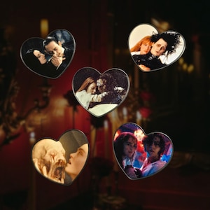 May include: Five heart-shaped pins featuring movie stills. The images include scenes from various films, with a focus on romantic or dramatic moments. The pins have a metallic border and are set against a dark background.