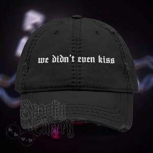 May include: Black distressed baseball cap with the text "we didn't even kiss" in white gothic font. The cap has a curved brim and a worn appearance. The brand "Spooky Cherry" is printed on the bottom.