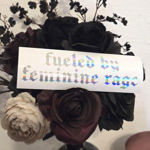May include: A bouquet of dark roses, including black, burgundy, and cream-colored blooms. A white rectangular sticker with the text "fueled by feminine rage" in a holographic, gothic font is placed in the center of the bouquet.
