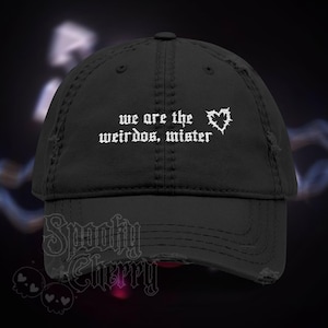 May include: Black distressed baseball cap with the text "we are the weirdos, mister" in white gothic font, along with a white heart graphic. The cap has a curved brim and a worn appearance, suggesting a vintage or edgy style.