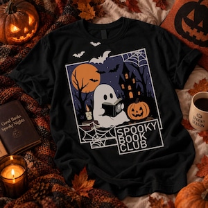 Può includere: Una t-shirt nera con una stampa grafica di un fantasma bianco che legge un libro, una zucca, una casa stregata, pipistrelli e una ragnatela. Il testo "SPOOKY BOOK CLUB" è stampato in bianco all'interno di una cornice rettangolare a destra.