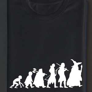 May include: Black t-shirt featuring a white silhouette design depicting the evolution of fantasy characters. The design includes figures of a hobbit, dwarf, warrior, elf, swordsman, and wizard.