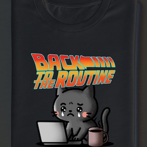 Op de afbeelding: Zwart T-shirt met een cartoonkat die huilt voor een laptop en een koffiemok. De tekst "BACK TO THE ROUTINE" staat in oranje en blauw. De kat is grijs met roze wangen.