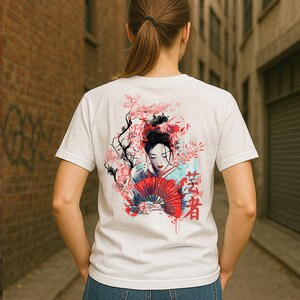 Può includere: T-shirt bianca con un disegno ad acquerello di una geisha con un ventaglio rosso, fiori di ciliegio e testo giapponese. L'opera d'arte utilizza tonalità di rosso, rosa, nero e blu. La maglietta è a girocollo classico.