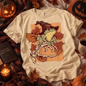 Può includere: Una t-shirt color crema con una stampa grafica di una rana verde che indossa un cappello da strega marrone scuro, appollaiata su una mano scheletrica circondata da foglie autunnali arancioni e marroni su sfondo rettangolare arancione.