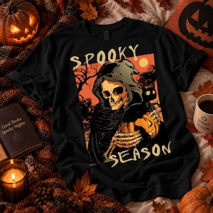 Può includere: Una t-shirt nera con una stampa grafica di uno scheletro incappucciato che tiene in mano una zucca e un gatto nero su sfondo arancione. Il testo "SPOOKY SEASON" è stampato in color crema sopra e sotto l'illustrazione centrale.