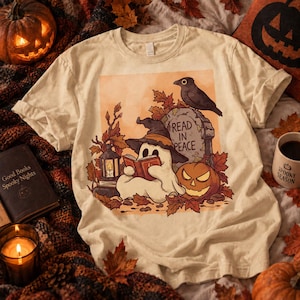 Può includere: Una t-shirt color crema con la grafica di un fantasma che indossa un cappello da strega e legge un libro. L'illustrazione include una lapide con il testo READ IN PEACE, un uccello nero, una zucca, una lanterna e foglie autunnali.