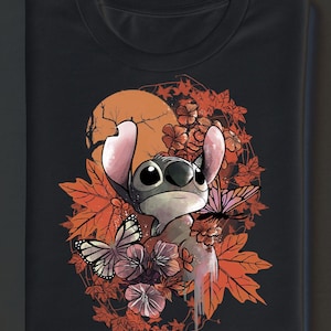 Può includere: T-shirt nero con un personaggio dei cartoni animati circondato da foglie autunnali, fiori e farfalle. Il design include una luna piena sullo sfondo. Il personaggio ha grandi occhi e orecchie rosa. Il design ha un effetto acquerello.
