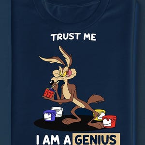 May include: A navy blue t-shirt with a cartoon Wile E. Coyote character holding a Rubik's Cube. The text "TRUST ME" is above the character, and "I AM A GENIUS" is below. Paint buckets in blue, white, yellow, and red are at the bottom.