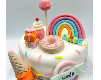 May include: A white cake decorated with a rainbow, a donut, a cupcake, a lollipop, a present, an ice cream cone, and sprinkles. The cake is decorated with the word "Sweetie".