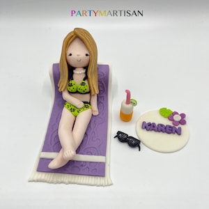 May include: A fondant cake topper featuring a woman in a green bikini lying on a purple beach towel. The topper also includes a cocktail, sunglasses, and a round cake topper with the name "KAREN" in purple letters.