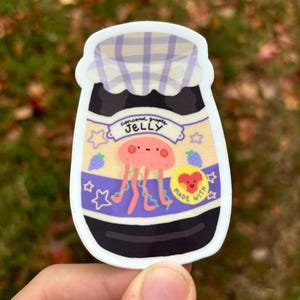 May include: A sticker of a glass jar with a purple and white checked lid. The jar is filled with a dark purple liquid and has a label that says "Concord Grape Jelly" with a cartoon jellyfish inside. The label also says "Made with Love".
