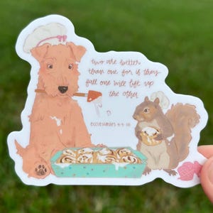 May include: A die-cut sticker featuring a tan dog and a brown squirrel wearing white chef hats. The dog holds a spatula over a teal baking dish of cinnamon rolls. Text reads: two are better than one for if they fall one will lift up the other.