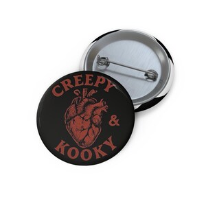 Könnte beinhalten: Zwei runde schwarze Ansteckbuttons mit Metallnadeln. Ein Button zeigt eine rote anatomische Herzillustration und die Worte "Creepy & Kooky" in Rot. Der andere Button ist silberfarben mit einem schwarz-weiß gestreiften Rand.