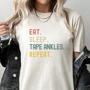 May include: A beige t-shirt with the text "EAT. SLEEP. TAPE ANKLES. REPEAT." printed in a vintage style font with red, yellow, and blue colors.