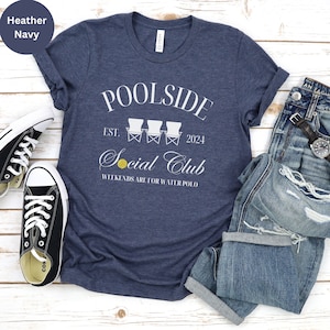 Pode incluir: Uma camiseta azul marinho heather com um design gr&aacute;fico branco que diz "Poolside Social Club Est. 2024" com uma ilustra&ccedil;&atilde;o branca de tr&ecirc;s cadeiras de praia e uma bola de v&ocirc;lei amarela. O texto abaixo do gr&aacute;fico diz "Weekends are for water polo."