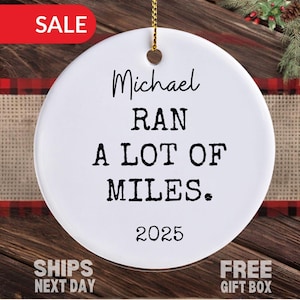May include: White ceramic ornament with the text "Michael RAN A LOT OF MILES. 2025" in black. The ornament has a gold string for hanging. A red "SALE" banner is in the top left corner.