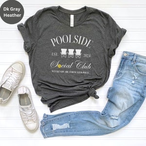 Pode incluir: Uma camiseta cinza escuro com um design gr&aacute;fico branco que diz "POOLSIDE Social Club EST. 2024" com tr&ecirc;s cadeiras de praia brancas e uma bola de v&ocirc;lei amarela. O texto abaixo das cadeiras diz "WEEKENDS ARE FOR WATER POLO".