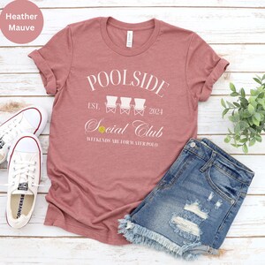 Pode incluir: Camiseta heather malva com texto branco que diz "Poolside Est. 2024 Social Club Weekends are for Water Polo". O texto &eacute; cercado por um gr&aacute;fico de tr&ecirc;s cadeiras de praia brancas com uma bola de v&ocirc;lei amarela.