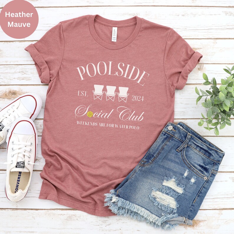Pode incluir: Camiseta heather malva com texto branco que diz "Poolside Est. 2024 Social Club Weekends are for Water Polo". O texto &eacute; cercado por um gr&aacute;fico de tr&ecirc;s cadeiras de praia brancas com uma bola de v&ocirc;lei amarela.