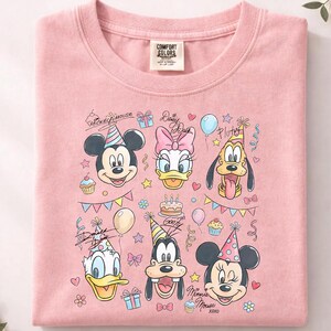 May include: A light pink t-shirt featuring a colorful graphic of Mickey Mouse, Daisy Duck, Pluto, Donald Duck, Goofy, and Minnie Mouse, all wearing party hats. The design includes birthday cake, balloons, and confetti.
