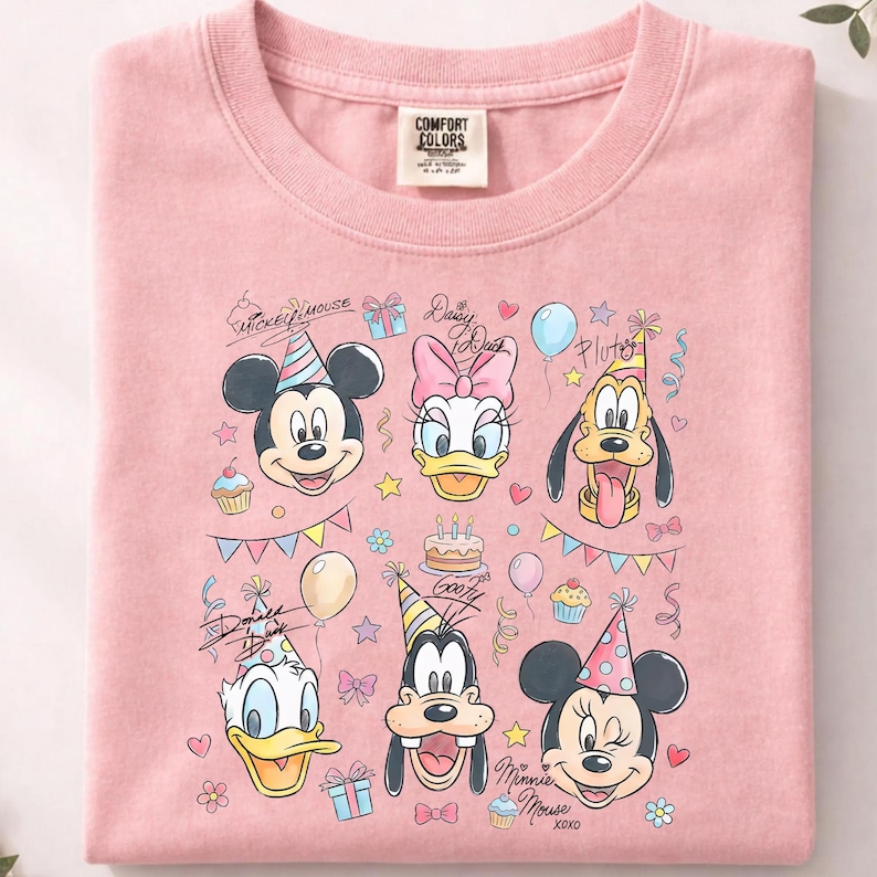 May include: A light pink t-shirt featuring a colorful graphic of Mickey Mouse, Daisy Duck, Pluto, Donald Duck, Goofy, and Minnie Mouse, all wearing party hats. The design includes birthday cake, balloons, and confetti.