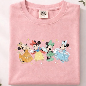 Op de afbeelding: Zachtroze t-shirt met een afbeelding van vier Minnie Mouse-personages in Disney-prinsessenkostuums. Minnie wordt afgebeeld als Belle in een gele jurk, Sneeuwwitje, een fee en Assepoester in een blauwe jurk. Het shirt heeft een Comfort Colors-label.