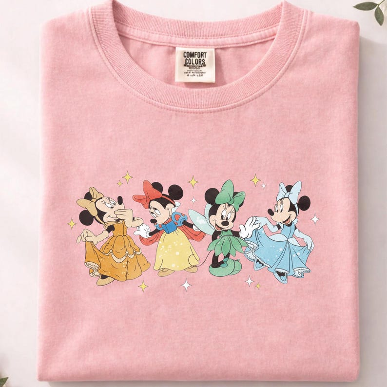 Op de afbeelding: Zachtroze t-shirt met een afbeelding van vier Minnie Mouse-personages in Disney-prinsessenkostuums. Minnie wordt afgebeeld als Belle in een gele jurk, Sneeuwwitje, een fee en Assepoester in een blauwe jurk. Het shirt heeft een Comfort Colors-label.
