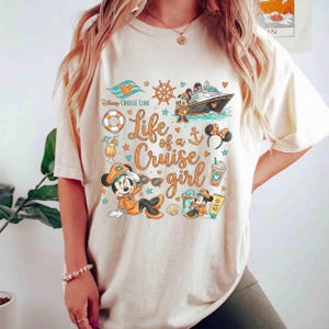 May include: Cream-colored t-shirt featuring a graphic design with Mickey Mouse in cruise attire, a cruise ship, and the text "Life of a Cruise Girl." The design includes nautical elements like a ship's wheel, anchor, and life preserver.