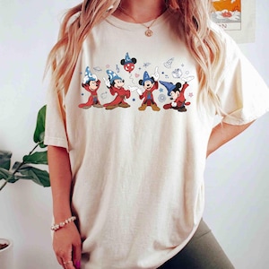 Könnte beinhalten: Cremefarbenes T-Shirt mit einer Grafik von Mickey Mouse in Zaubererkleidung. Die Zeichentrickfiguren Mickey Mouse tragen rote und blaue Roben und Hüte mit Sternen und Noten. Das Shirt hat eine lockere Passform.