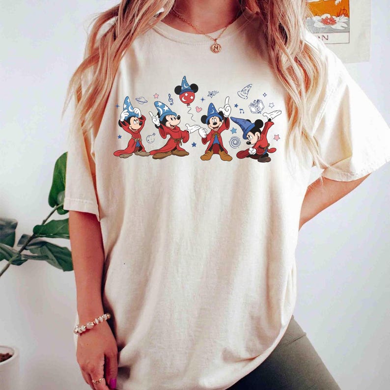 May include: Cream-colored t-shirt featuring a graphic of Mickey Mouse in wizard attire. The cartoon Mickey Mouse characters are in red and blue robes and hats, with stars and musical notes. The shirt is a relaxed fit.