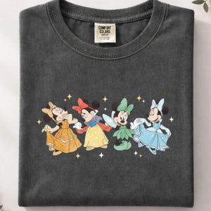 Op de afbeelding: Donkergrijs Comfort Colors T-shirt met een afbeelding van vier Minnie Mouse-personages in prinsessenoutfits. De afbeelding bevat gele, rode, groene en blauwe jurken met steraccenten. Het shirt heeft een ronde hals en korte mouwen.