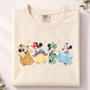 Op de afbeelding: Cr&egrave;mekleurig Comfort Colors T-shirt met vier Minnie Mouse-personages in prinsessenoutfits. De afbeelding toont Minnie in een gele jurk, ge&iuml;nspireerd op Sneeuwwitje, een fee en Assepoester, met sterren.