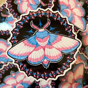 Puede incluir: Una pegatina con una polilla colorida con alas rosas, azules y blancas. La polilla está rodeada por un diseño circular negro y rosa.