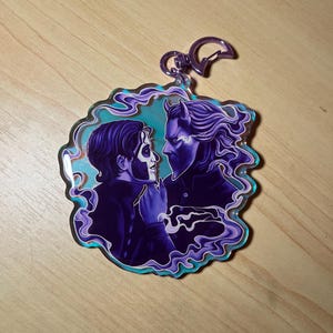 May include: An acrylic keychain featuring a purple-toned illustration of two figures facing each other, one with face paint and the other with horns, surrounded by swirling purple smoke and a light blue background, attached to a purple metal clasp.