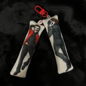 May include: Two white rectangular keychains with images of a person in black and white face paint and black clothing. One keychain has a red jacket. The keychains are attached to a red clasp.