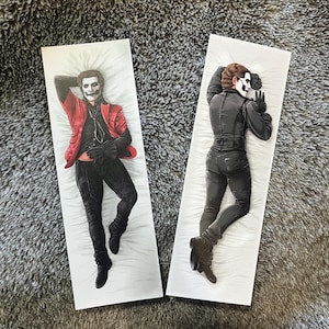 May include: Two body pillow covers featuring illustrations of a figure in a theatrical costume. One cover shows the figure in a red jacket, black pants, and gloves. The other shows the figure in a black outfit. Both are on white pillow backgrounds.