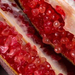May include: Close-up of a halved pomegranate, showcasing vibrant red seeds and a white membrane. The seeds are numerous, round, and juicy, filling the fruit's interior. The outer skin is a light tan color.