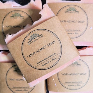 May include: Four bars of pink soap with brown paper labels. The labels have a circle with the text "OUTDOOR VERB" and "ANTI-AGING" SOAP. The text below the circle says "Rosemary, Rose, Rosehip Oil" and "handmade in Texas".