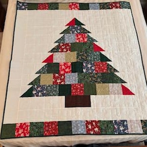 May include: A handmade Christmas tree quilt with a white background. The tree is constructed from various fabric squares in shades of green, red, and blue. The quilt has a dark green border and a matching border at the top and bottom.