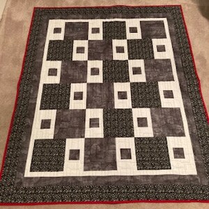 May include: A quilt with a geometric pattern of squares in shades of gray and white. The quilt has a red border.