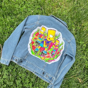 May include: A blue denim jacket with a colourful embroidered patch on the back. The patch features three cartoon characters skateboarding in a fiery design.