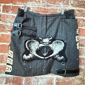 Puede incluir: Falda gris a rayas finas con detalles de hebillas de cuero negro y un gran gráfico anatómico de pelvis en blanco y negro. La falda está colgada en una percha transparente contra una pared de ladrillos.