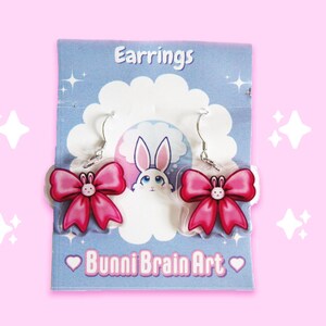 May include: A pair of clear acrylic earrings featuring a pink bow design with a small bunny face in the center. The earrings hang from silver hooks and are displayed on a blue card with the text "Earrings" and "Bunni Brain Art".