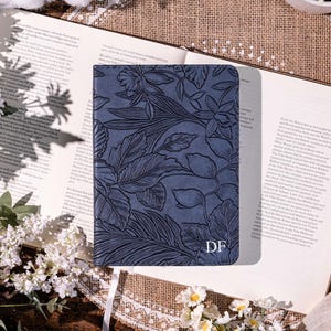 May include: A navy blue journal with a floral design and the initials "DF" is open on a book. The journal is surrounded by white flowers, cotton, and greenery. The book's pages are visible, and the scene is lit by natural light.