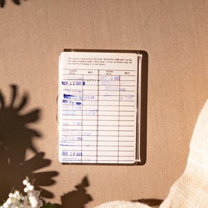 May include: A vintage library card with handwritten entries and stamps, resting on a tan surface. The card is surrounded by white daisies and a cream-colored fabric, with shadows adding depth. The text on the card is legible.