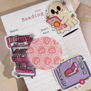 May include: A collection of stickers and a reading log. The stickers include a pink strawberry-patterned pop socket, a ghost holding a book, a bookshelf, and a card that says "WORLD: OFF" and "BOOK: ON". The reading log is labeled "2025 Reading".