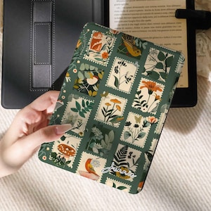 以下が含まれることがあります： 黒いKindleデバイスと、緑と白の花柄のKindleケース。ケースは、植物のイラストが描かれた切手のような正方形のデザインが特徴です。Kindleは開いており、テキストが表示されています。